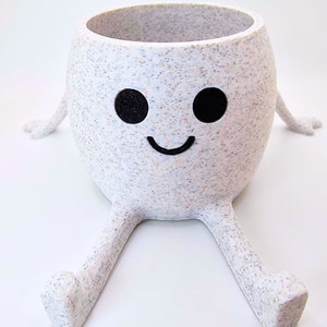 May include: White ceramic planter with a speckled finish shaped like a smiling face with two black eyes and outstretched arms and legs.