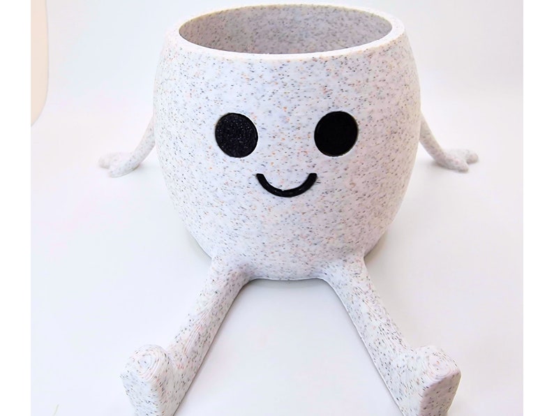 May include: White ceramic planter with a speckled finish shaped like a smiling face with two black eyes and outstretched arms and legs.