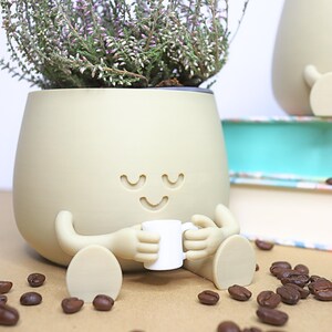 May include: A beige ceramic planter shaped like a person with a smiling face and closed eyes. The planter is holding a small white cup and is sitting on a brown surface with coffee beans scattered around it.