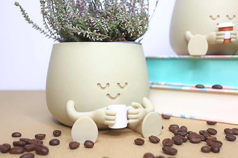May include: A beige ceramic planter shaped like a person with a smiling face and closed eyes. The planter is holding a small white cup and is sitting on a brown surface with coffee beans scattered around it.