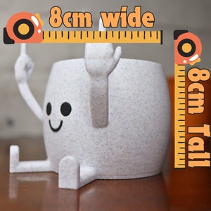 May include: A white ceramic planter with a smiling face and arms and legs. The planter is 8 cm wide and 8 cm tall.