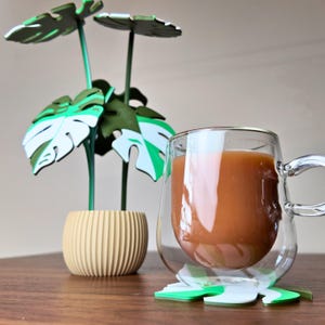 May include: A clear glass mug filled with a brown beverage, resting on a leaf-shaped coaster. A decorative plant with green and white leaves and a beige pot is to the left. The mug has a handle and a double-walled design.