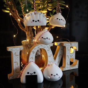 May include: Five white ceramic ornaments shaped like dumplings and a rice ball, with cute faces and black eyes. The ornaments are hanging from red and white strings and are arranged in front of a wooden "LOVE" sign with a string of lights.