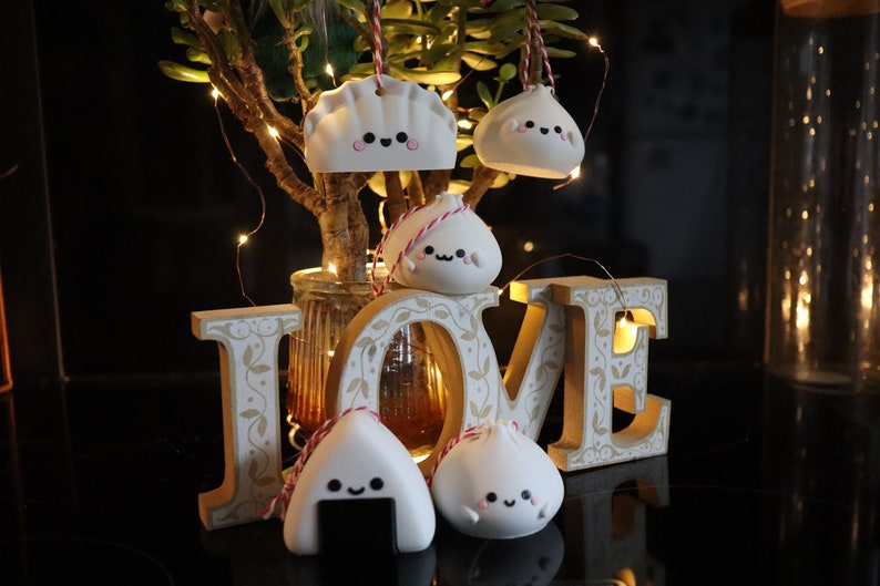 May include: Five white ceramic ornaments shaped like dumplings and a rice ball, with cute faces and black eyes. The ornaments are hanging from red and white strings and are arranged in front of a wooden "LOVE" sign with a string of lights.