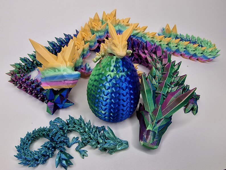 May include: A set of 3D printed iridescent dragon figurines. The dragons are in various poses, including a curled up dragon, a dragon with its wings spread, and a dragon with its head raised. The dragons are all made of a shiny, iridescent material that changes color in the light.