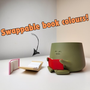 Puede incluir: Un macetero de cer&aacute;mica verde con forma de persona con una cara sonriente, sosteniendo un libro rojo. Dos libros est&aacute;n sobre la mesa, uno es rosa y el otro beige. El texto "Swappable book colours!" est&aacute; escrito sobre el macetero.