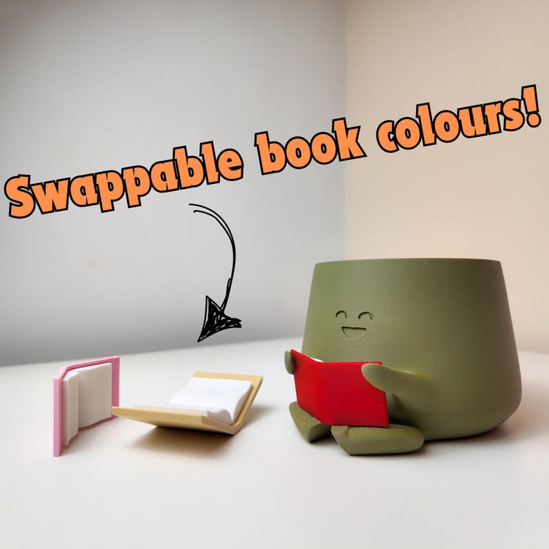 Puede incluir: Un macetero de cer&aacute;mica verde con forma de persona con una cara sonriente, sosteniendo un libro rojo. Dos libros est&aacute;n sobre la mesa, uno es rosa y el otro beige. El texto "Swappable book colours!" est&aacute; escrito sobre el macetero.
