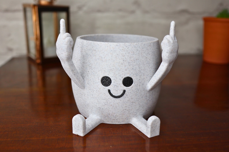 May include: A white ceramic planter shaped like a person with two arms raised in a middle finger gesture. The planter has a smiling face with black eyes and a black mouth.