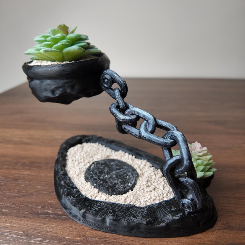 May include: A black planter with a chain design, holding two small succulents. The base is avocado-shaped, filled with sand and a central black circle. The chain supports a floating pot with a green succulent.