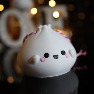 May include: White ceramic dumpling ornament with a pink and white string, black eyes, and a pink smile. The ornament is sitting on a black surface.