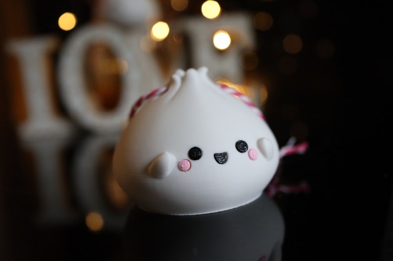 May include: White ceramic dumpling ornament with a pink and white string, black eyes, and a pink smile. The ornament is sitting on a black surface.