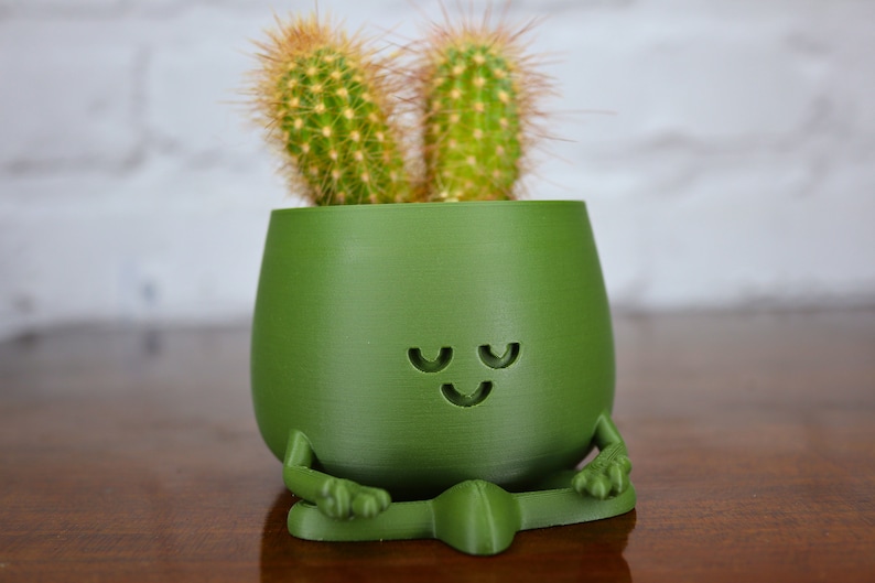 Puede incluir: Una maceta de cer&aacute;mica verde con forma de figura meditando con dos cactus creciendo dentro. La maceta tiene una cara sonriente y los ojos cerrados.