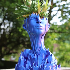 May include: A blue decorative planter in the shape of a human head and neck, with a succulent plant growing from the top. The planter has a unique design with a dripping effect, and is made of a solid material.