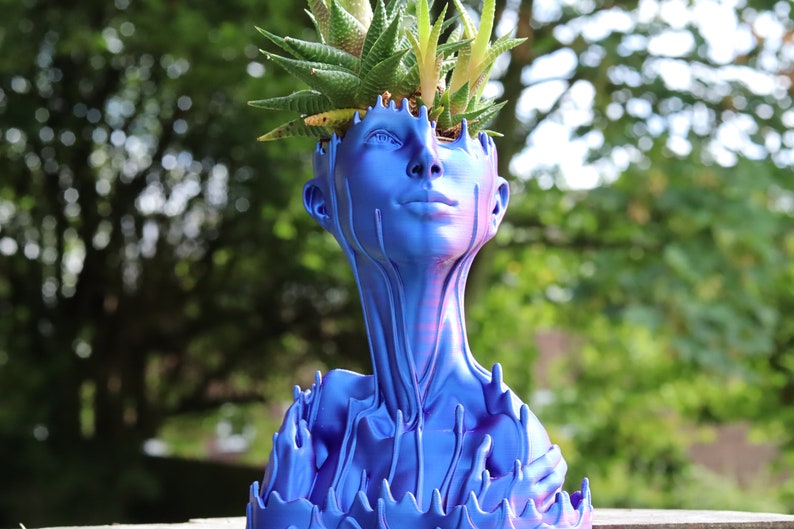 May include: A blue decorative planter in the shape of a human head and neck, with a succulent plant growing from the top. The planter has a unique design with a dripping effect, and is made of a solid material.