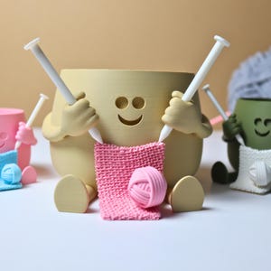May include: Three ceramic yarn bowl holders with smiling faces, each holding knitting needles and a ball of yarn. The bowls are pink, beige, and green.