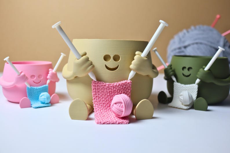 May include: Three ceramic yarn bowl holders with smiling faces, each holding knitting needles and a ball of yarn. The bowls are pink, beige, and green.