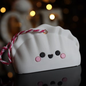 May include: White ceramic ornament shaped like a dumpling with a pink and white string attached. The dumpling has a smiling face with black eyes and a pink blush.
