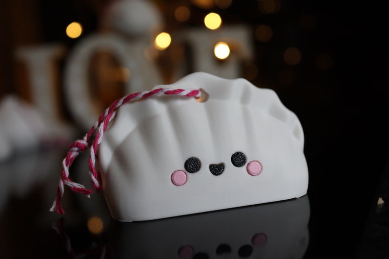 May include: White ceramic ornament shaped like a dumpling with a pink and white string attached. The dumpling has a smiling face with black eyes and a pink blush.