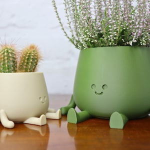 Puede incluir: Dos macetas de cer&aacute;mica con forma de personajes sonrientes con piernas y brazos. La maceta m&aacute;s peque&ntilde;a es beige y contiene un cactus. La maceta m&aacute;s grande es verde y contiene una planta con flores.