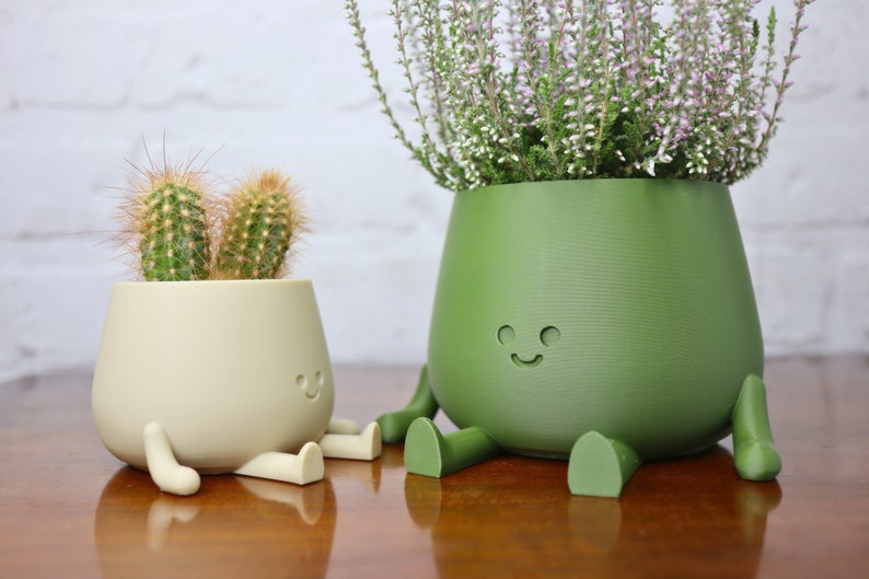 Puede incluir: Dos macetas de cer&aacute;mica con forma de personajes sonrientes con piernas y brazos. La maceta m&aacute;s peque&ntilde;a es beige y contiene un cactus. La maceta m&aacute;s grande es verde y contiene una planta con flores.
