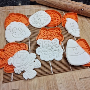 May include: A collection of Halloween-themed cookie cutters and cookies. The cutters are orange, and the cookies are white, featuring designs like a jack-o'-lantern, ghost, mummy, and candy corn. The words "Trick or Treat" are also visible.