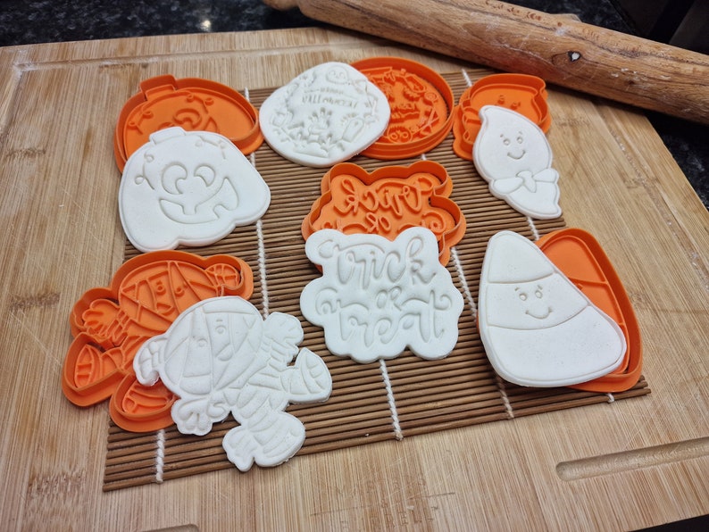 May include: A collection of Halloween-themed cookie cutters and cookies. The cutters are orange, and the cookies are white, featuring designs like a jack-o'-lantern, ghost, mummy, and candy corn. The words "Trick or Treat" are also visible.