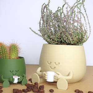 May include: Two green and beige ceramic planters shaped like cartoon characters with smiling faces. The planters are holding a cactus and a flowering plant. The planters have small arms and legs and are holding tiny coffee cups.