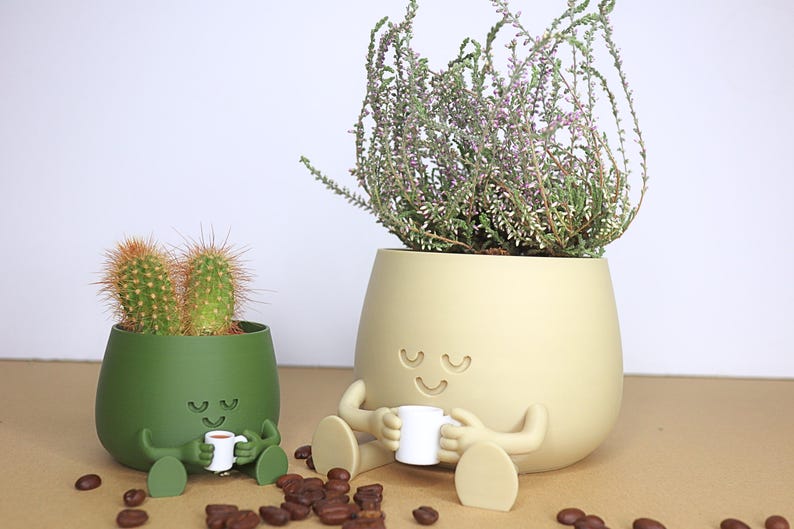 May include: Two green and beige ceramic planters shaped like cartoon characters with smiling faces. The planters are holding a cactus and a flowering plant. The planters have small arms and legs and are holding tiny coffee cups.