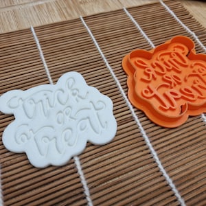 May include: Two cookie cutters, one white and one orange, sit on a bamboo mat. The white cutter has the words "Trick or Treat" in a cursive font. The orange cutter is the same shape and has the same text.