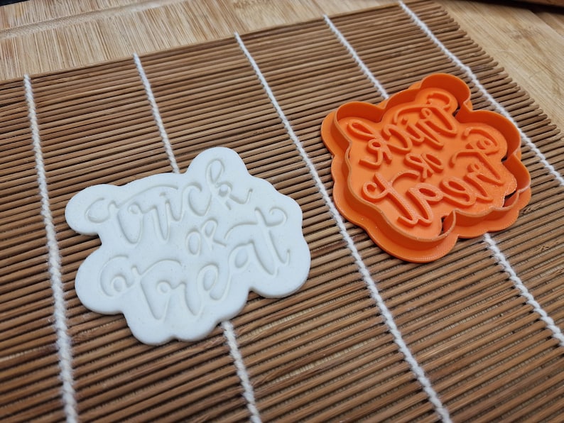 May include: Two cookie cutters, one white and one orange, sit on a bamboo mat. The white cutter has the words "Trick or Treat" in a cursive font. The orange cutter is the same shape and has the same text.
