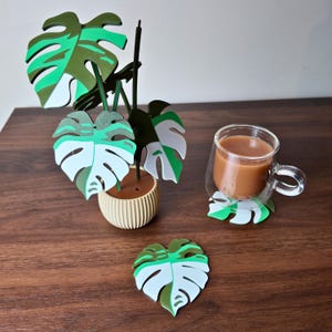 May include: A decorative set featuring a small artificial plant and leaf-shaped coasters. The plant has green and white leaves in a beige pot. A glass mug with coffee sits on a matching coaster. A single coaster is also visible.