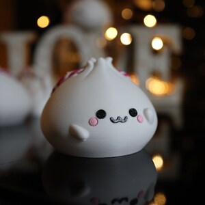 May include: White ceramic figurine shaped like a dumpling with a pink and red design, black eyes, and a black mustache. The figurine has a cute, cartoon-like expression.