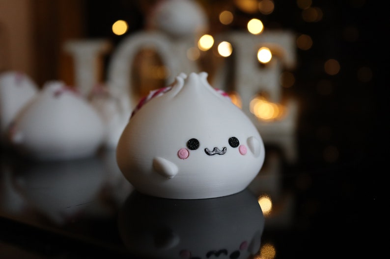 May include: White ceramic figurine shaped like a dumpling with a pink and red design, black eyes, and a black mustache. The figurine has a cute, cartoon-like expression.