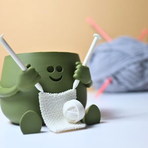 May include: A green ceramic yarn bowl shaped like a smiling face with arms and legs, holding knitting needles and a ball of yarn.