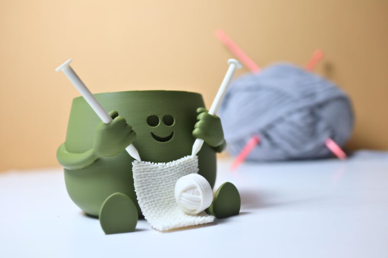May include: A green ceramic yarn bowl shaped like a smiling face with arms and legs, holding knitting needles and a ball of yarn.