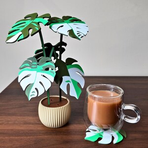 May include: A decorative plant with green, white, and black leaves in a beige pot, next to a glass mug of coffee on a matching leaf-shaped coaster. The plant has a modern, minimalist design.