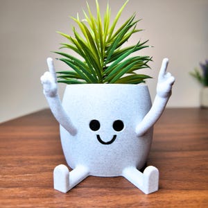 Puede incluir: Una maceta blanca caprichosa con una cara sonriente, brazos levantados y piernas extendidas. La maceta contiene una planta verde y amarilla vibrante. El dise&ntilde;o es l&uacute;dico y decorativo, adecuado para uso en interiores.