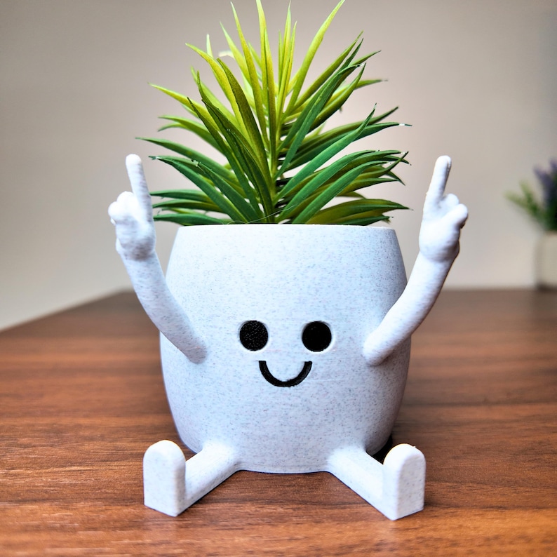 Puede incluir: Una maceta blanca caprichosa con una cara sonriente, brazos levantados y piernas extendidas. La maceta contiene una planta verde y amarilla vibrante. El dise&ntilde;o es l&uacute;dico y decorativo, adecuado para uso en interiores.