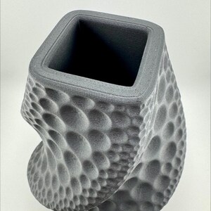 May include: A gray, textured vase with a square opening and a twisted, bubble-like pattern. The vase has a modern design and a matte finish, suitable for home decor.