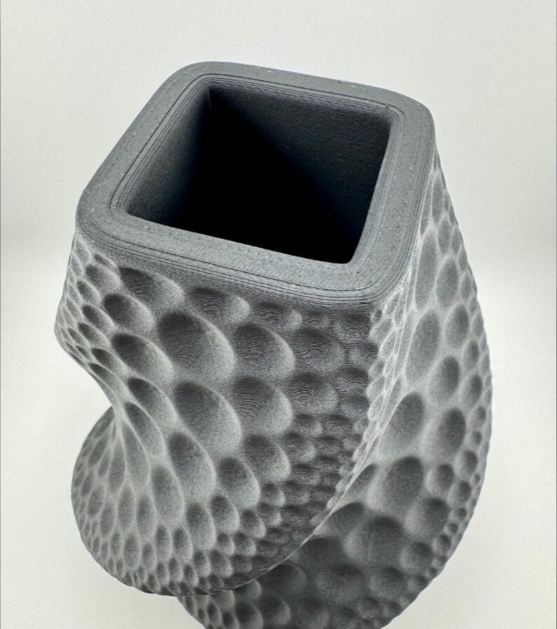 May include: A gray, textured vase with a square opening and a twisted, bubble-like pattern. The vase has a modern design and a matte finish, suitable for home decor.