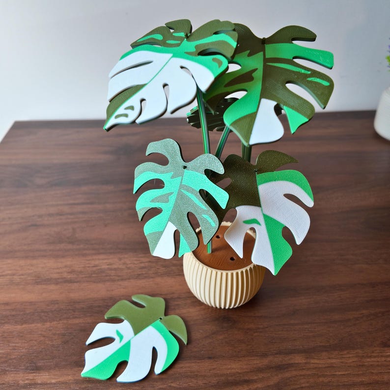 May include: A decorative artificial Monstera plant with green, white, and teal leaves. The plant is in a beige pot with a ribbed design. One leaf is separate from the plant. The leaves have a split-leaf design.