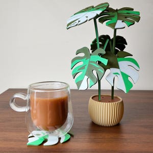May include: A clear glass mug with a handle, filled with a light brown beverage, sits on a leaf-shaped coaster. A decorative paper plant with green and white leaves is in a beige pot. The plant has green stems.