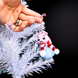May include: A pink pig ornament with a red and white scarf and a blue hat hanging on a white artificial Christmas tree.