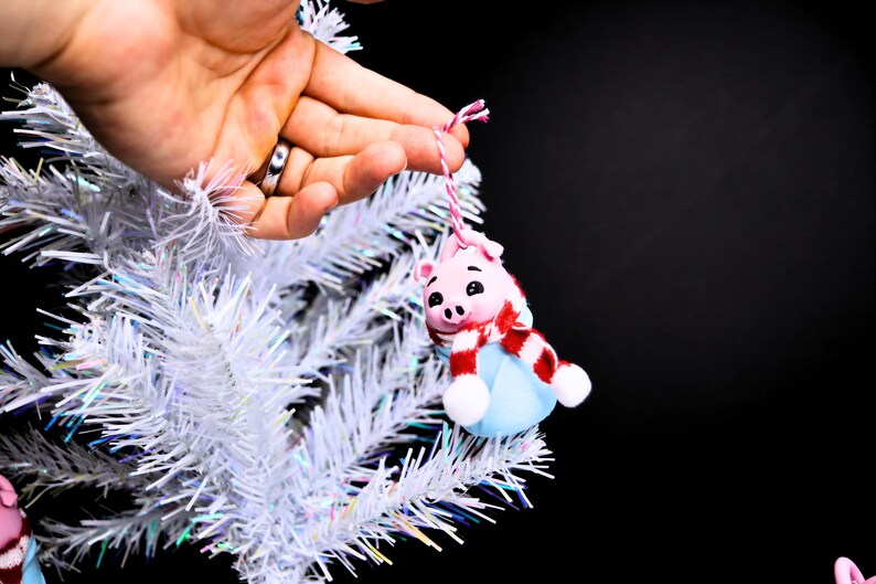 May include: A pink pig ornament with a red and white scarf and a blue hat hanging on a white artificial Christmas tree.
