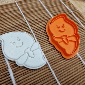 May include: A white ghost-shaped cookie cutter and a bright orange cutter sit on a bamboo mat. The ghost design has a smiling face and a bow tie. The cookie cutter is made of plastic and is likely used for baking.