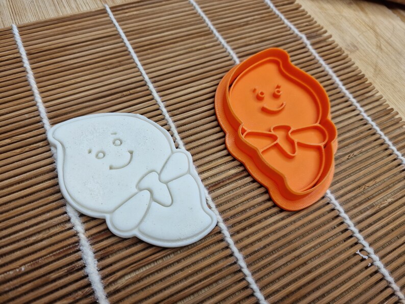 May include: A white ghost-shaped cookie cutter and a bright orange cutter sit on a bamboo mat. The ghost design has a smiling face and a bow tie. The cookie cutter is made of plastic and is likely used for baking.