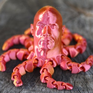 May include: A 3D printed sculpture of a human head with a red, metallic finish, attached to a red, segmented octopus body.