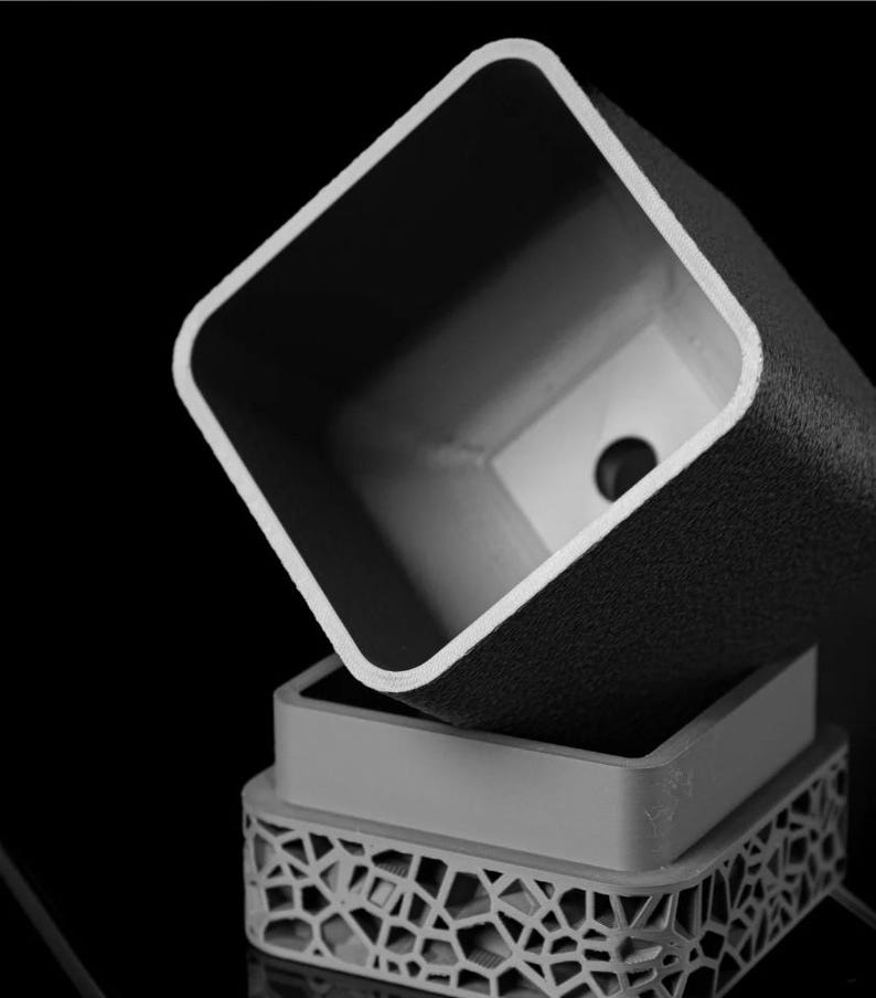 May include: A grayscale image of three stacked, square-shaped planters. The top planter is dark with a textured surface, the middle one is smooth and light, and the bottom one has a geometric, openwork design. The planters are likely for indoor or outdoor use.