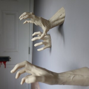 May include: Three realistic-looking, white, plaster hands reaching out from a wall. The hands are posed as if they are grabbing something.