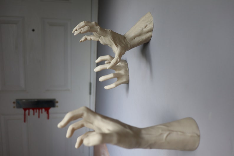 May include: Three realistic-looking, white, plaster hands reaching out from a wall. The hands are posed as if they are grabbing something.